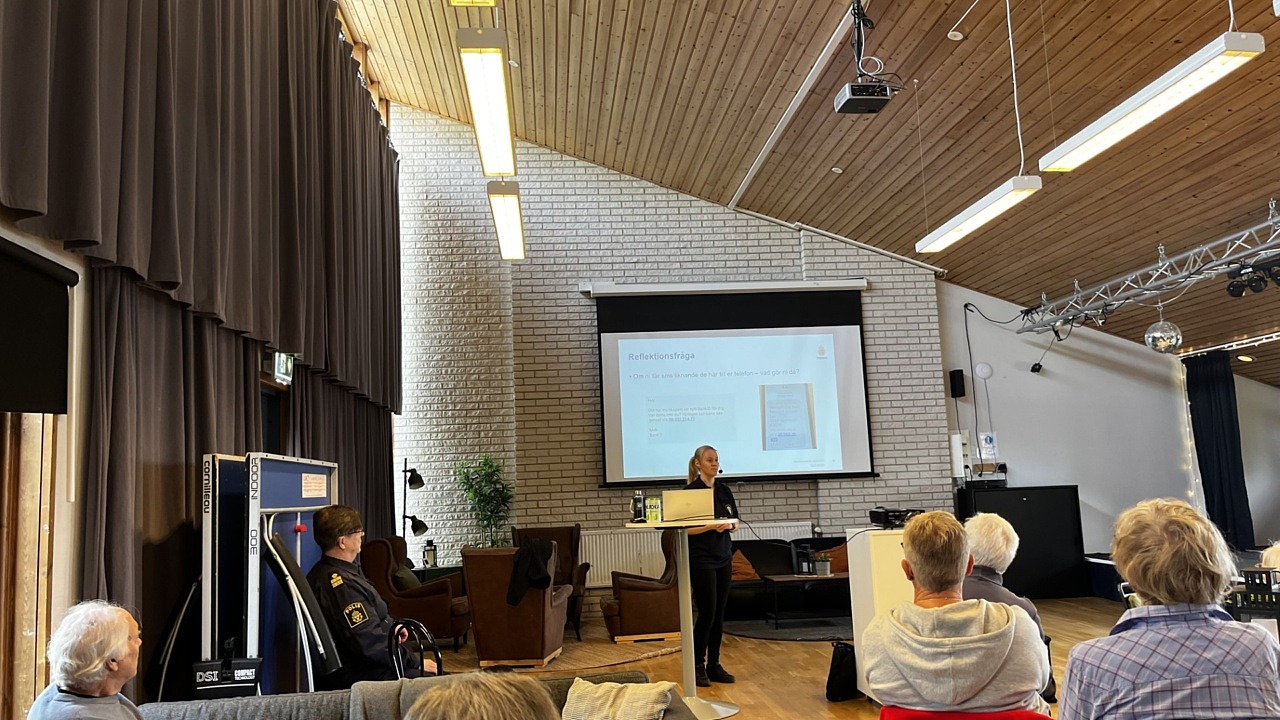 Människor som syns bakifrån, sittandes, lyssnar på föreläsare. Föreläsaren står upp och har sin presentation på en stor, vit duk.