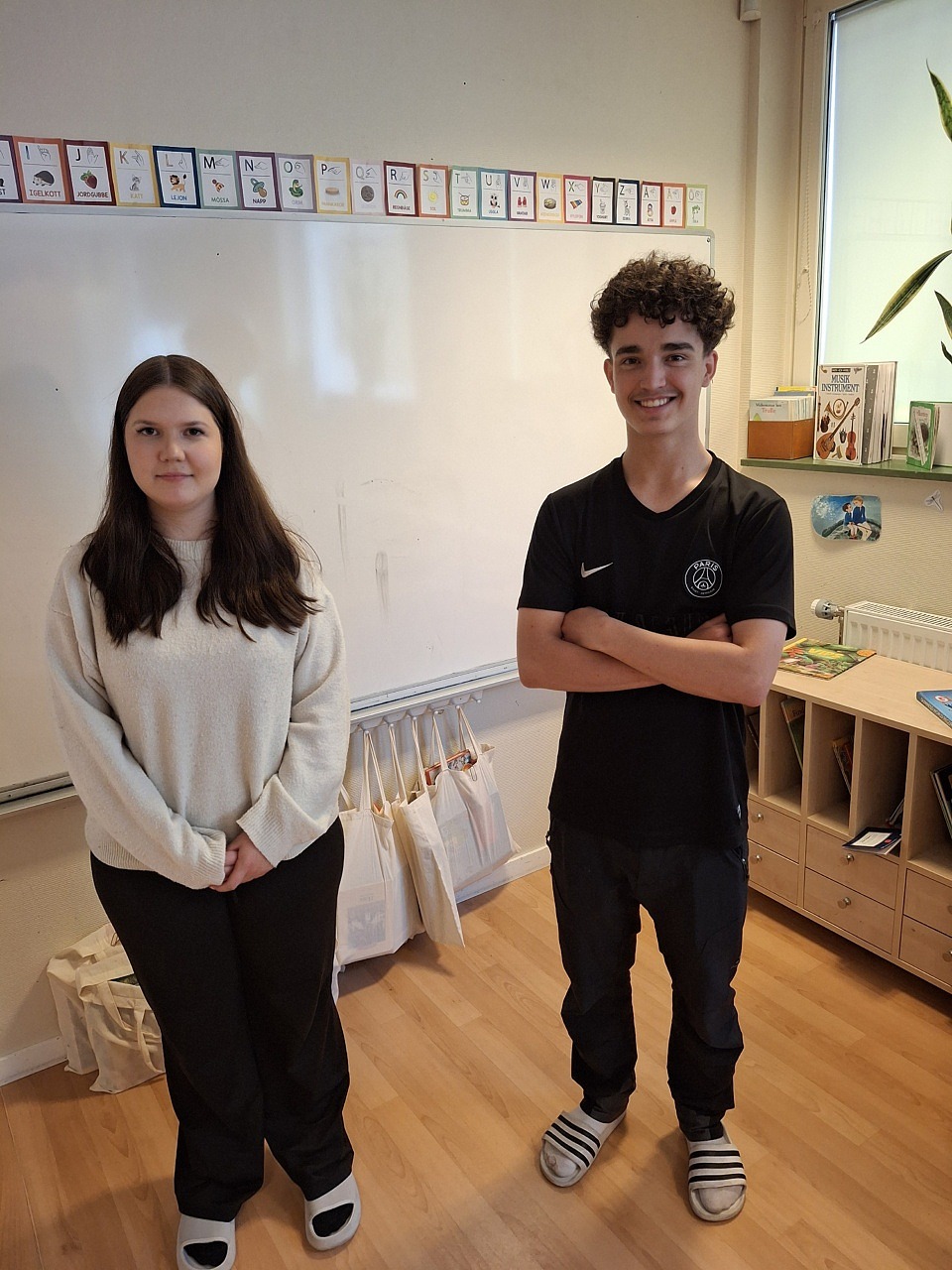 Emilia och Moaawia står och upp och tittar in i kameran. De ler. Bakom dem syns bilder på alfabetet och böcker.
