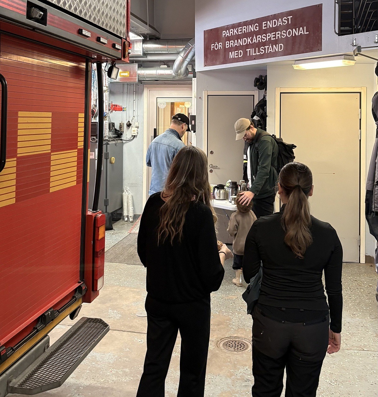 Inne på brandstationen cirkulerar människor i olika åldrar. Man ser ryggen på dem och bredvid dem står en röd brandbil.