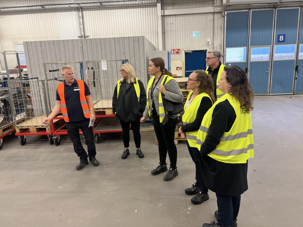 Fyra personer med gul väst står och lyssnar på en person i orange väst. Miljön runt dem är steril och grå.